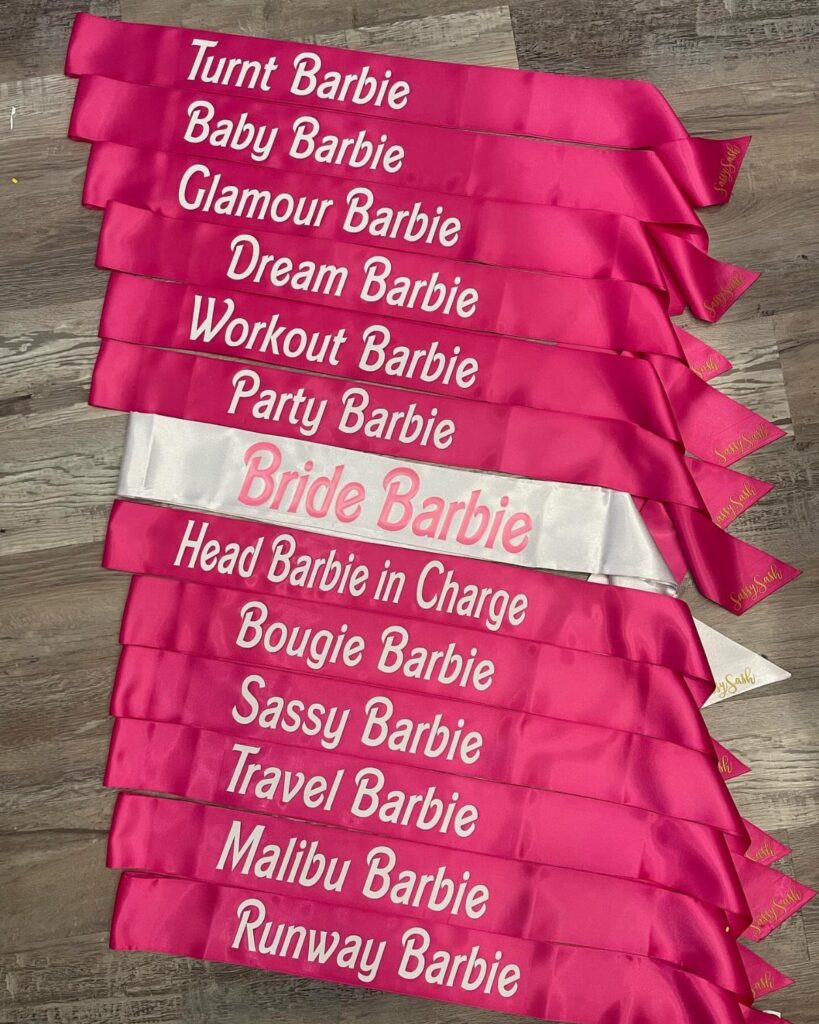 Pink and white satin sashes with Barbie themed text on a grey floor