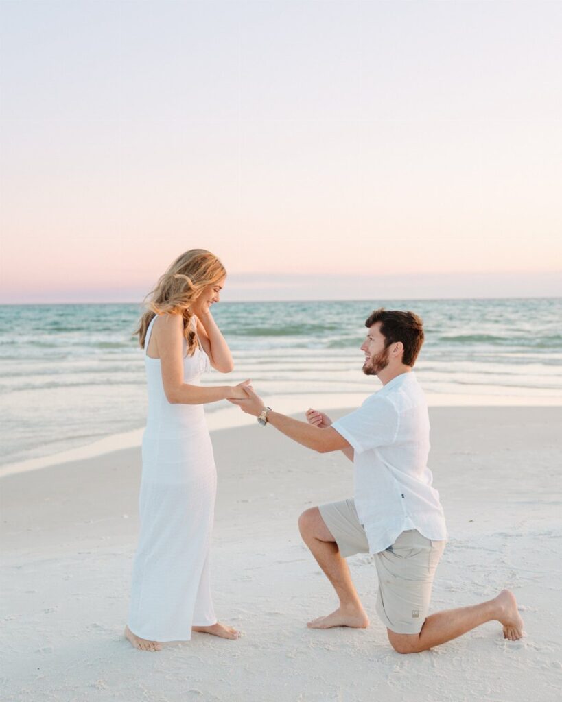 Man kneeling on a white sandy beach proposing to a woman in white.
