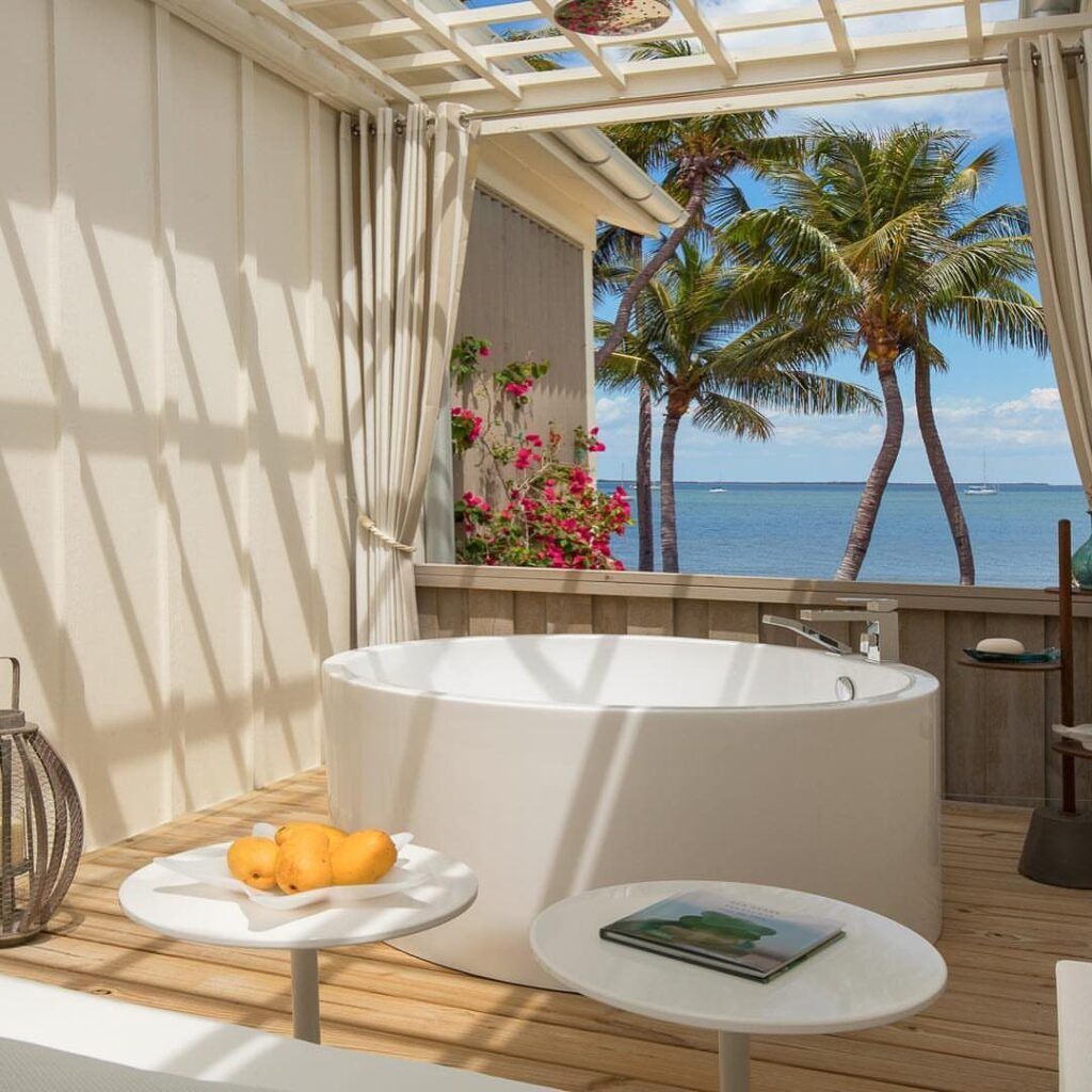 White freestanding bathtub on a wooden deck overlooking palm trees and blue ocean.