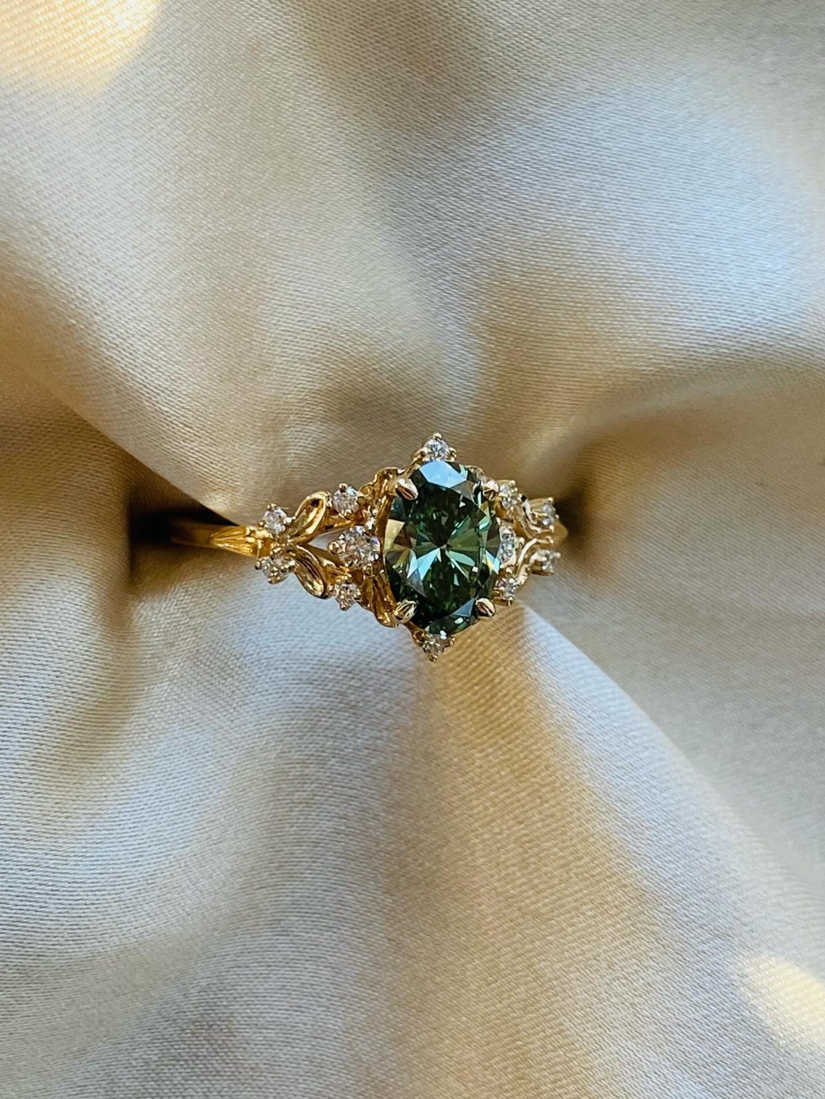 Gold engagement ring with oval green gemstone and leaf filigree on cream satin.