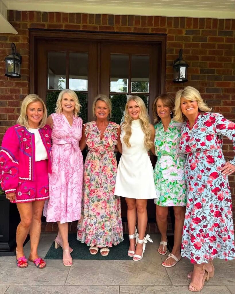 Six women posing in colorful floral dresses and outfits in front of a house.