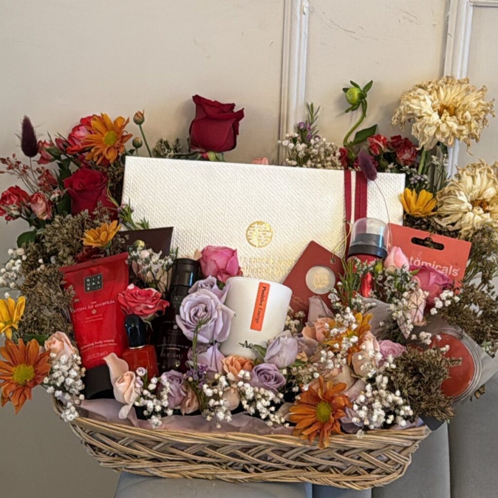 Wicker gift basket containing beauty products, a white box, and colorful fresh flowers.