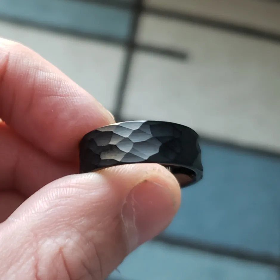 Black hammered wedding band held between a person's thumb and index finger.