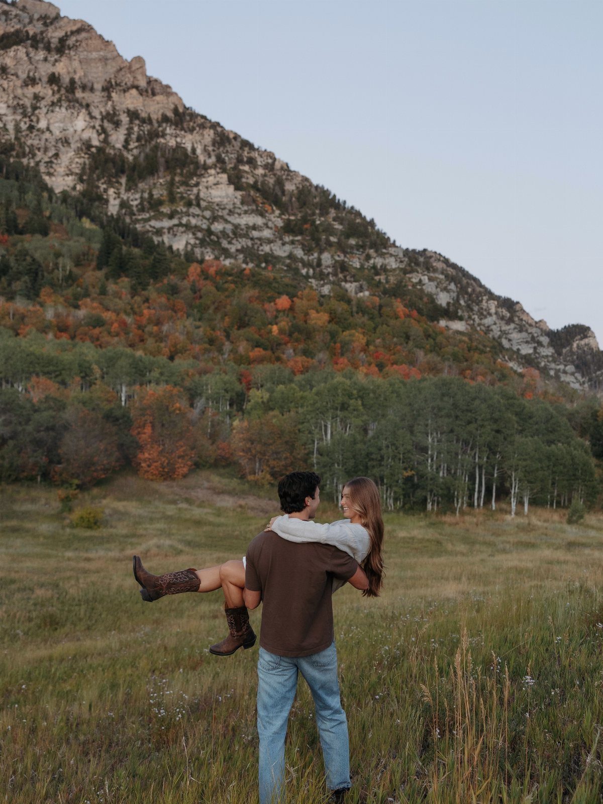 Couple embracing in a grassy field with autumn colored trees and mountains behind.
