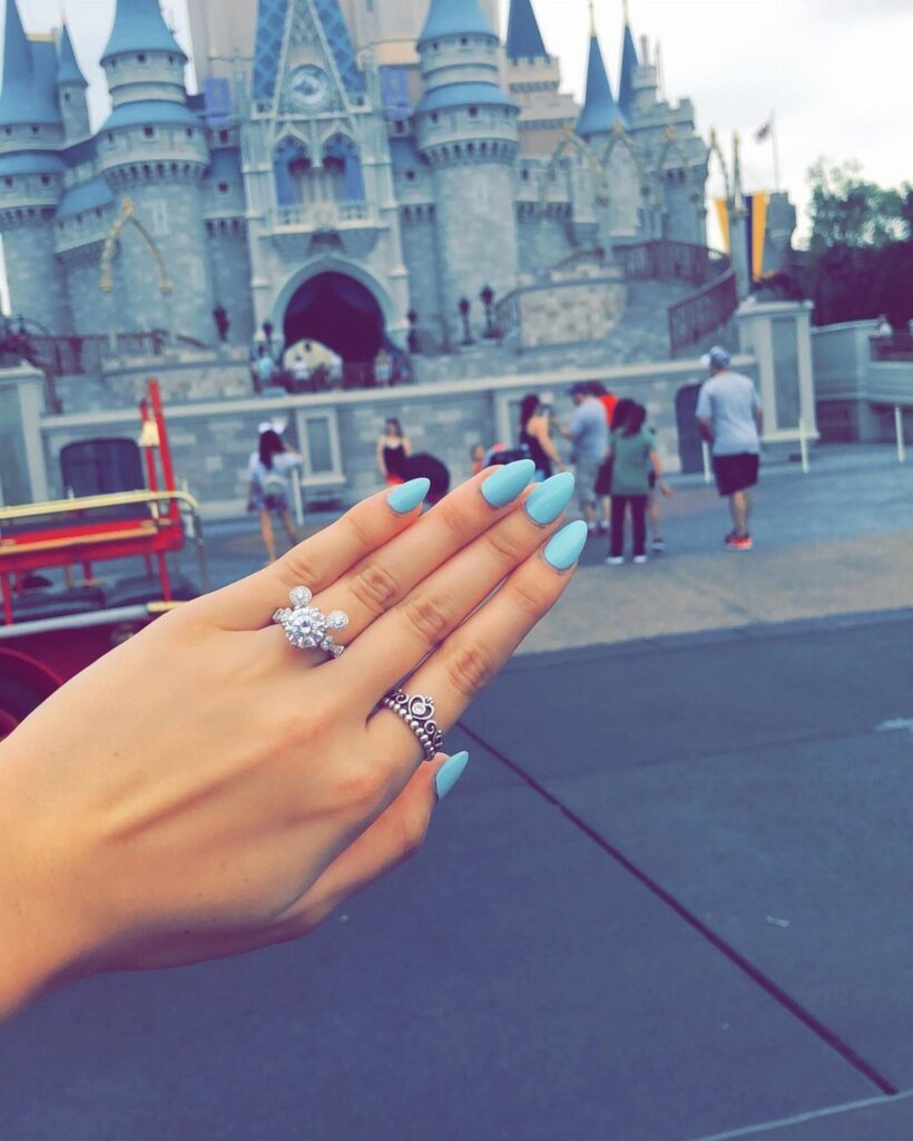 Mickey Mouse diamond engagement ring on a hand with blue nails before Cinderella Castle