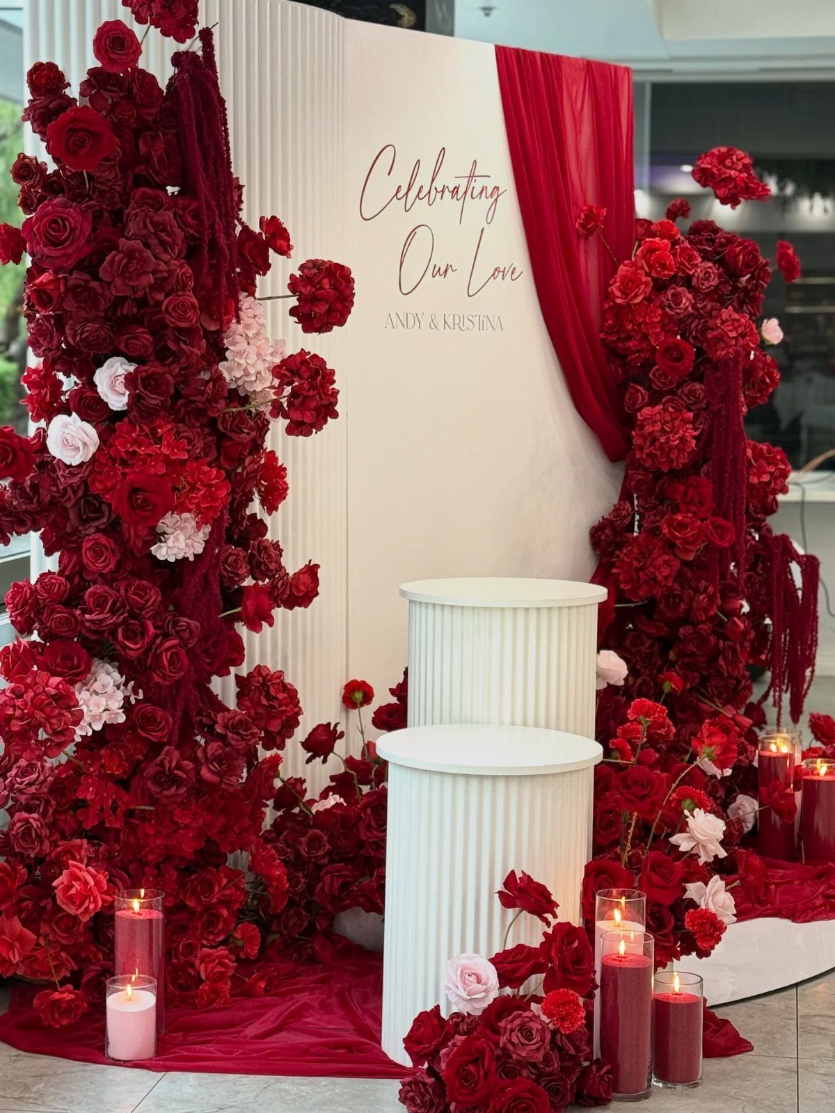 Red and pink rose floral backdrop with white pedestals and romantic event signage.
