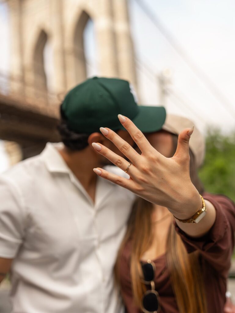 Woman's hand showing engagement ring with blurred couple kissing before Brooklyn Bridge