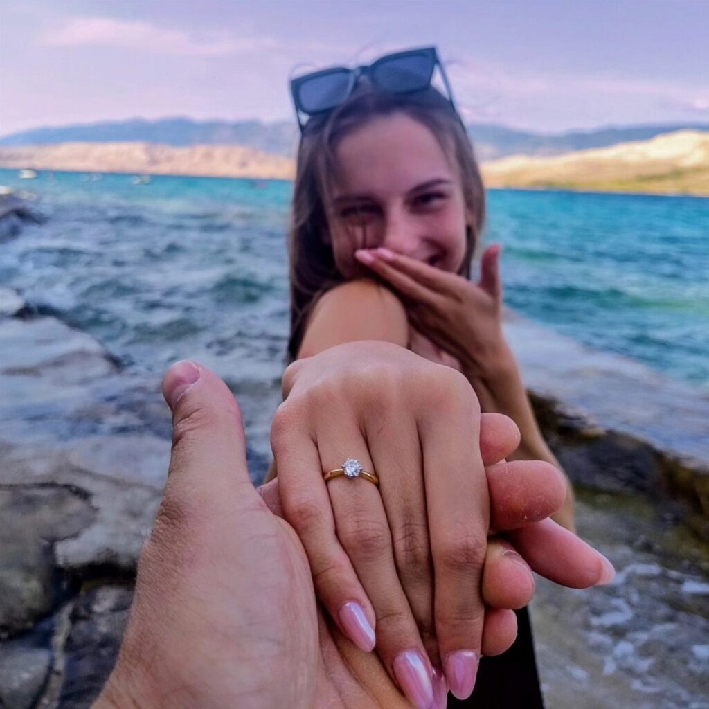 Hands holding with a diamond engagement ring visible against a scenic lake background.