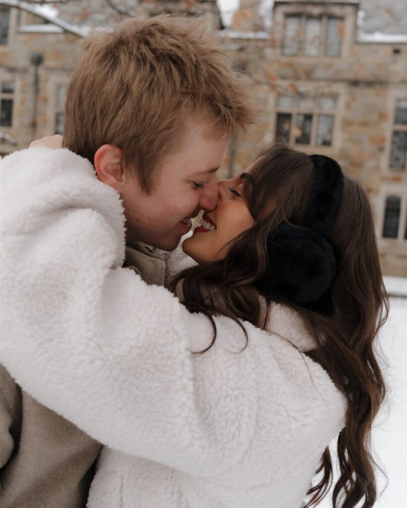 Couple embracing and smiling outdoors in winter attire against a stone building background.