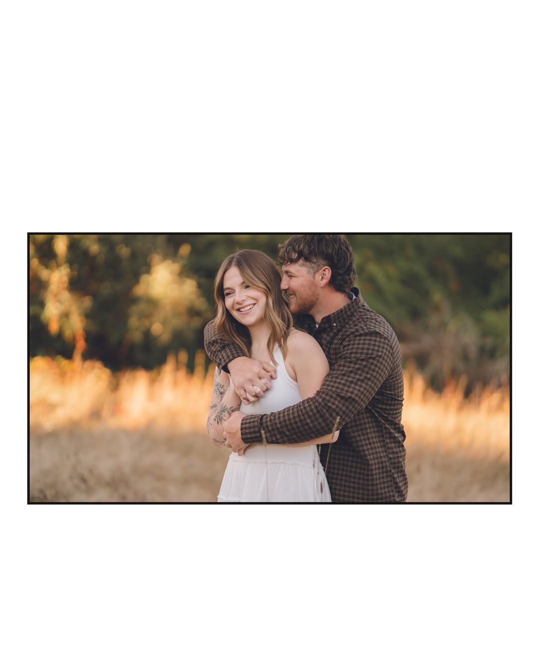 Couple embracing in a golden field, woman in white dress, man in plaid shirt.