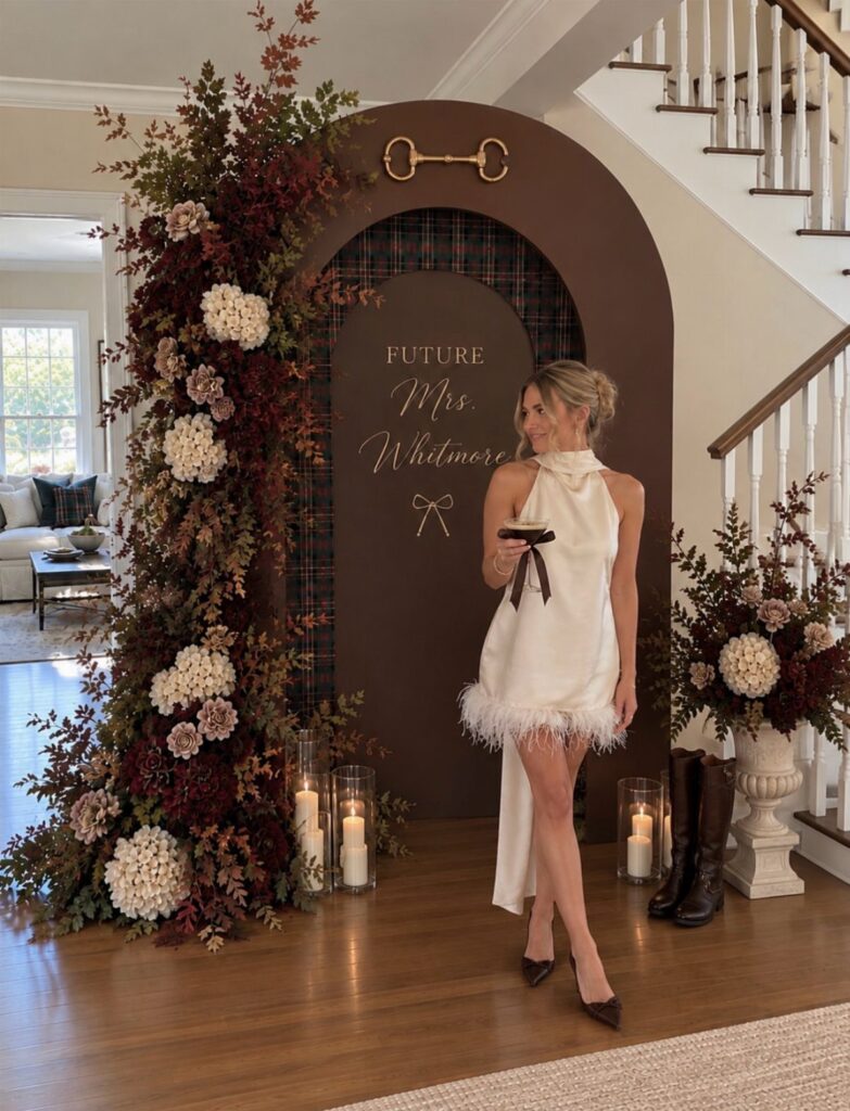 Woman in white dress posing by a fall-themed bridal shower backdrop with flowers.