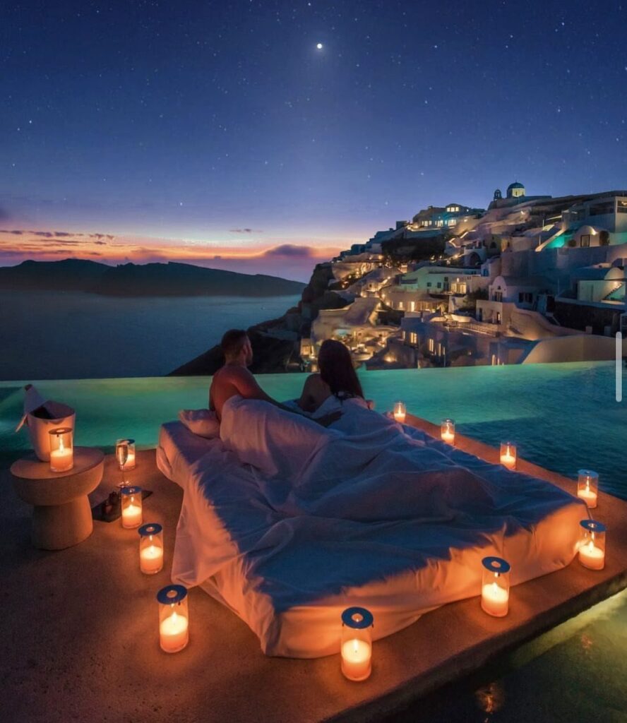 Couple in a candlelit outdoor bed overlooking the white buildings of Santorini at dusk.