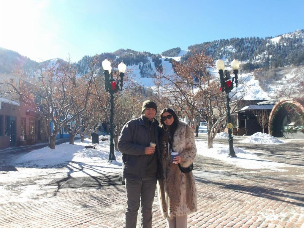 Couple in winter coats standing on a snowy street with mountains behind them.