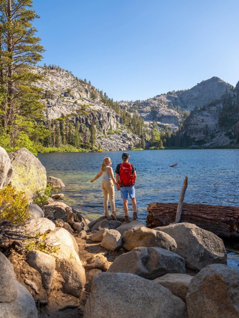Couple with red backpack standing by a clear alpine lake and granite mountains