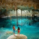 Couple holding hands overlooking a turquoise cenote inside a limestone cave in Mexico.