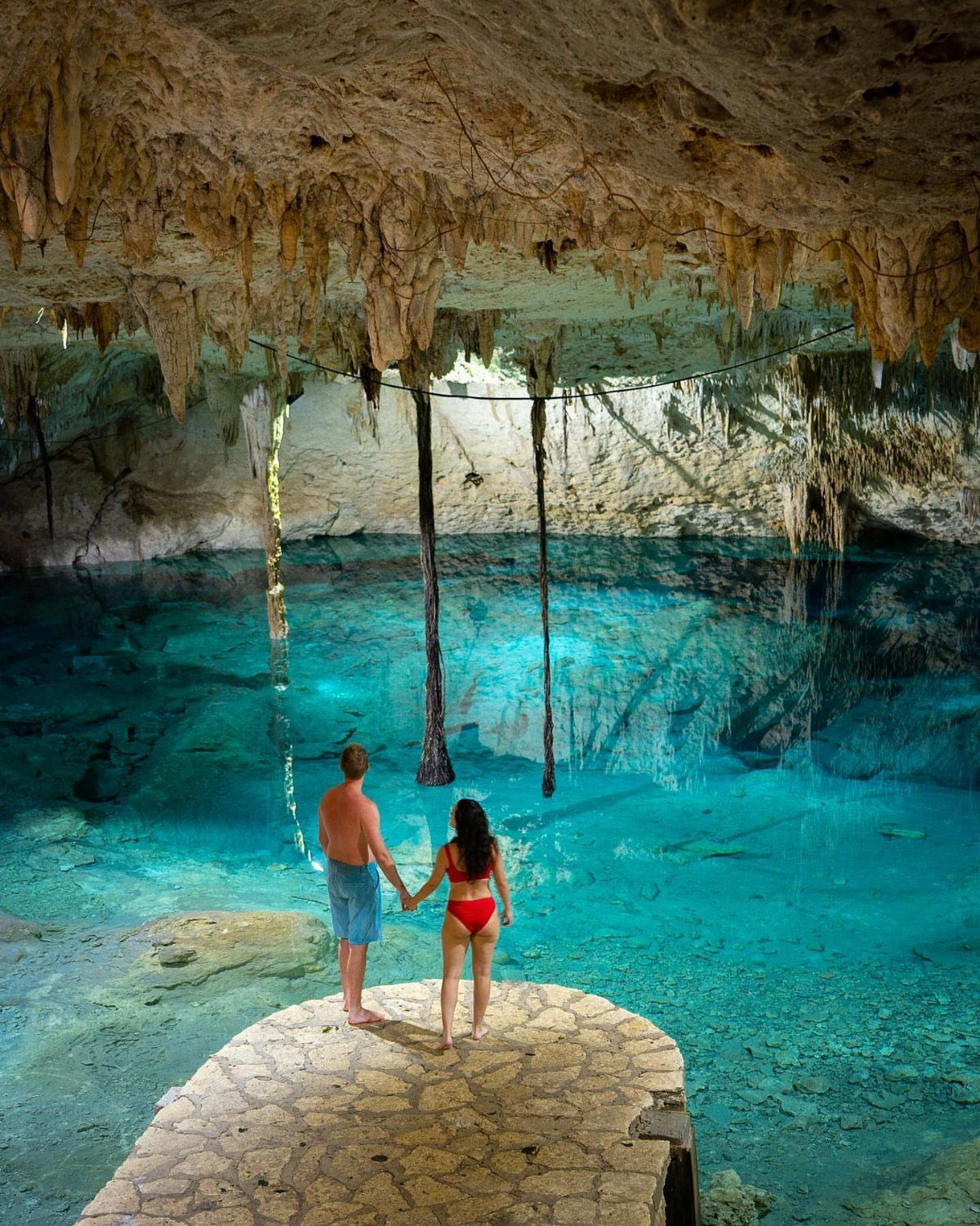 Couple holding hands overlooking a turquoise cenote inside a limestone cave in Mexico.