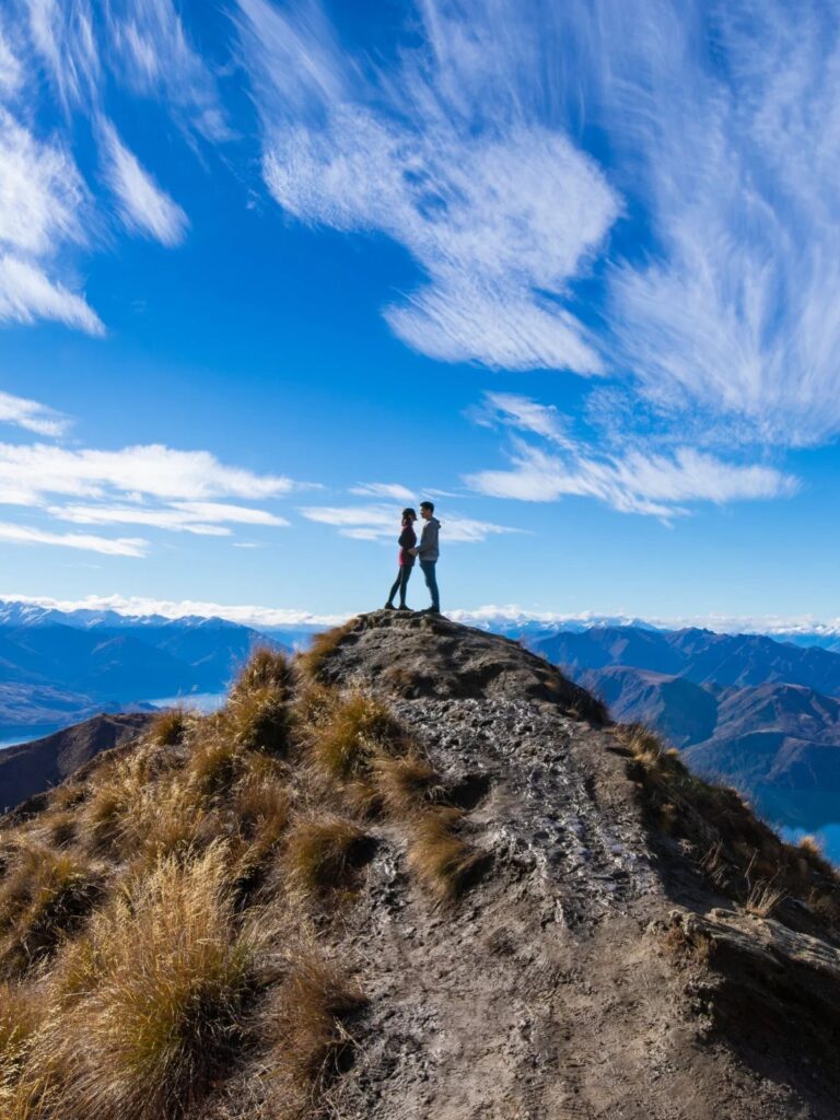 Couple standing on a mountain peak overlooking a vast range of blue mountains.