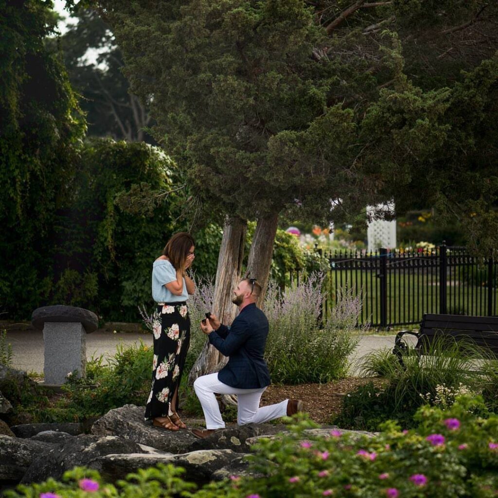 Man kneeling on a rocky garden path proposing to a woman in floral skirt