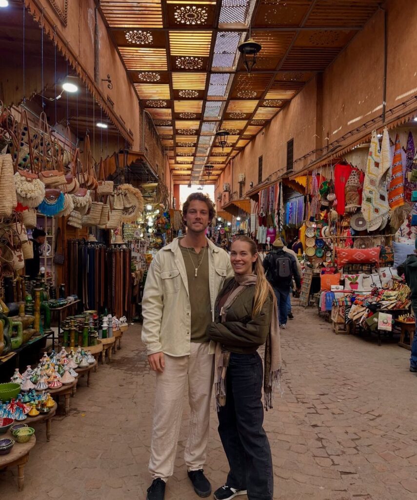 Couple smiling in a vibrant Marrakech market with colorful textiles and ceramics.