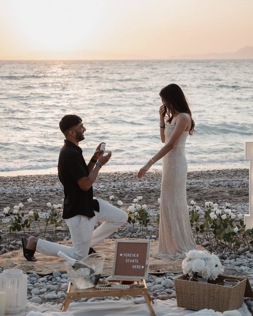Man proposing to a woman on a pebble beach with romantic floral decorations.