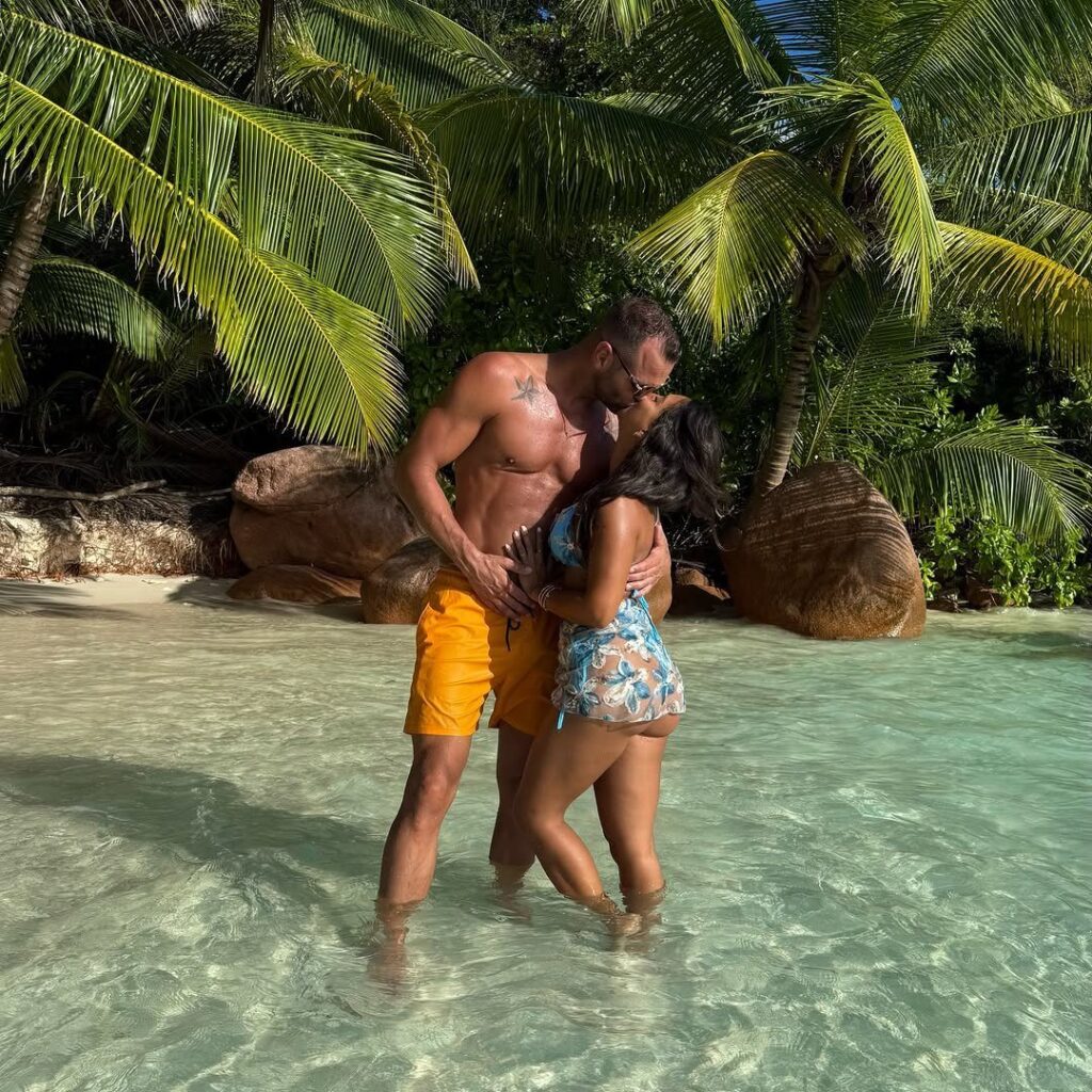 Couple embracing and kissing in turquoise water with palm trees and boulders.