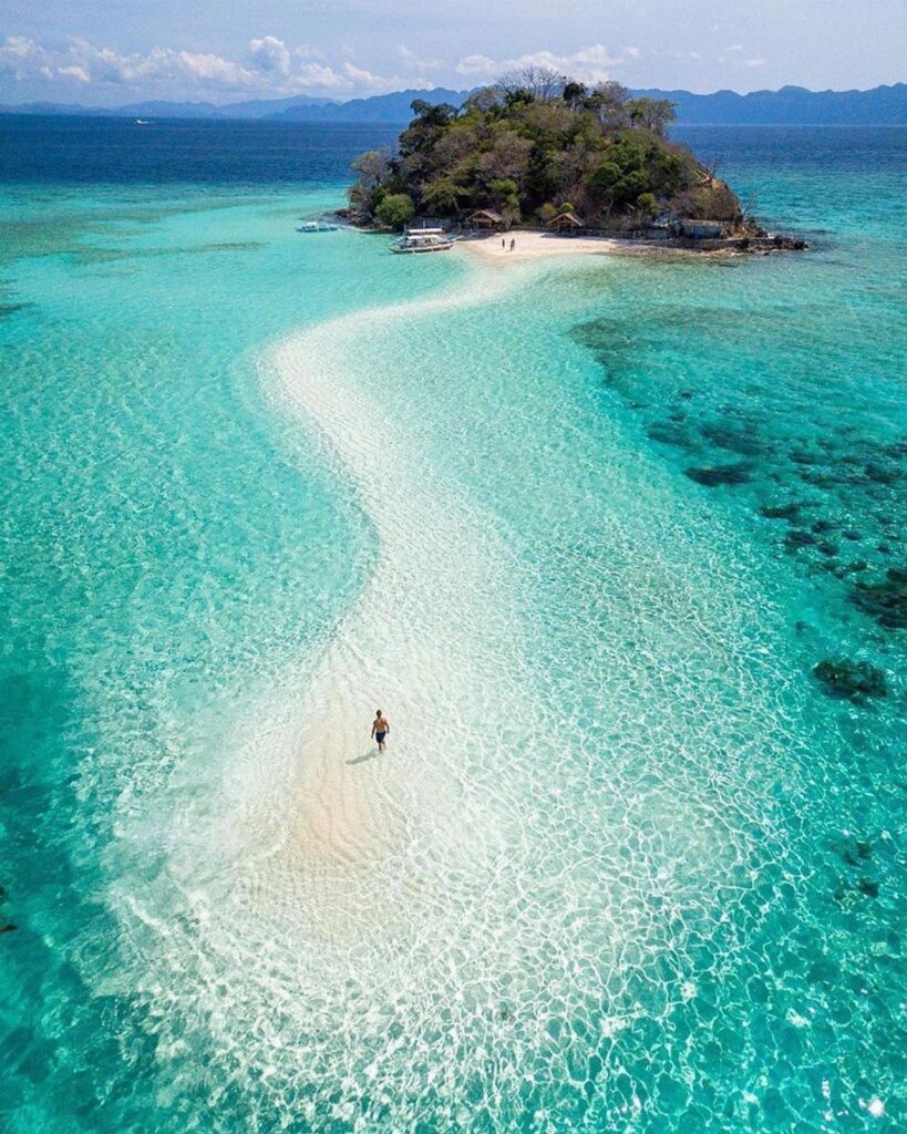 White sandbar curving through turquoise water toward a small tropical island.