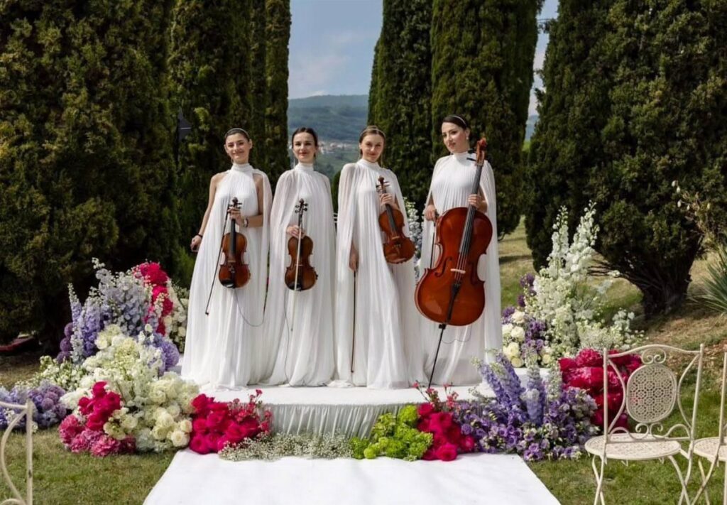 Four female string musicians in white gowns posing with instruments amidst wedding flowers.