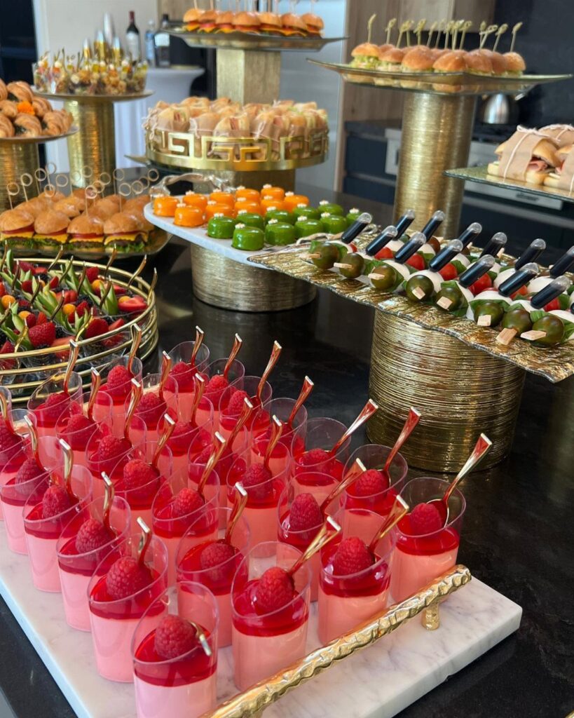 Gourmet catering spread with pink raspberry desserts and mini sliders on gold stands.